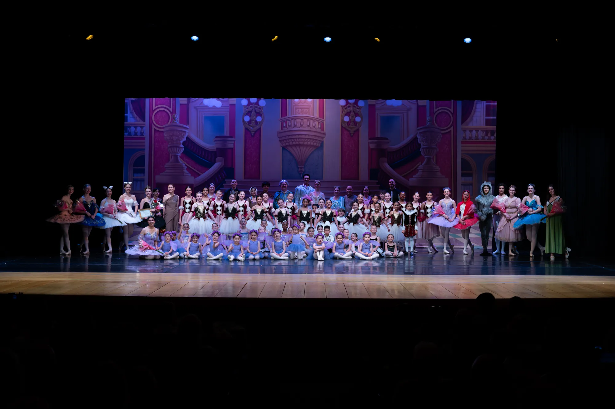 A large cast of dancers in colorful costumes lined up across a theater stage
